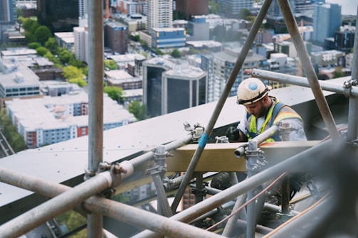 A construction worker wearing a hard hat and safety vest is working on a scaffolding high above a cityscape. The background shows a view of high-rise buildings surrounded by greenery.
