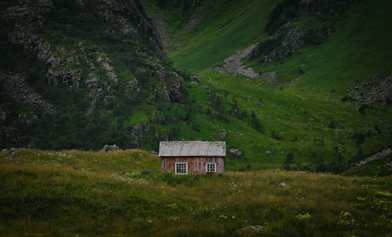 A solitary wooden cabin with a faded red exterior sits amidst a lush green valley. The landscape is dominated by towering hills covered in vibrant grass and scattered shrubs, creating a serene and secluded atmosphere.