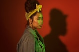 A person with a distinctive style stands in profile against a red background, casting a large shadow. They wear a denim jacket, large hoop earrings, and a yellow headband that ties up their hair. The lighting casts a green tint over the face and upper body, adding to the dramatic effect.