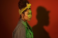 A person with a distinctive style stands in profile against a red background, casting a large shadow. They wear a denim jacket, large hoop earrings, and a yellow headband that ties up their hair. The lighting casts a green tint over the face and upper body, adding to the dramatic effect.
