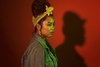A person with a distinctive style stands in profile against a red background, casting a large shadow. They wear a denim jacket, large hoop earrings, and a yellow headband that ties up their hair. The lighting casts a green tint over the face and upper body, adding to the dramatic effect.