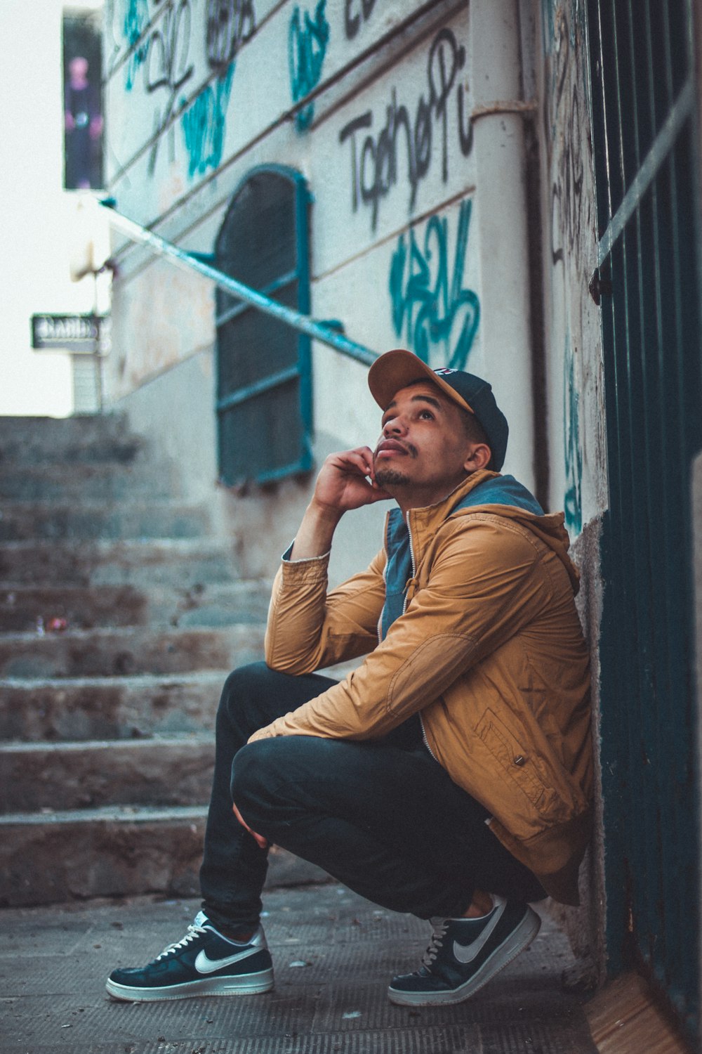 man wearing brown jacket sitting while leaning on wall at stair