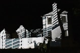 A building is illuminated in the dark with patterns of light and shadow creating an architectural illusion. The projected design features geometric lines and shapes, giving the façade a surreal and artistic appearance.