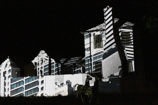 A building is illuminated in the dark with patterns of light and shadow creating an architectural illusion. The projected design features geometric lines and shapes, giving the façade a surreal and artistic appearance.
