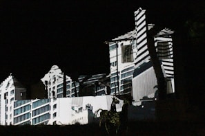 A building is illuminated in the dark with patterns of light and shadow creating an architectural illusion. The projected design features geometric lines and shapes, giving the façade a surreal and artistic appearance.