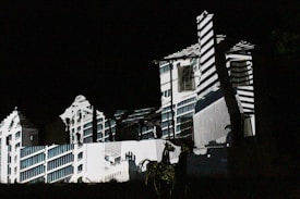 A building is illuminated in the dark with patterns of light and shadow creating an architectural illusion. The projected design features geometric lines and shapes, giving the fa&ccedil;ade a surreal and artistic appearance.