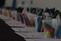 A row of plastic cups filled with colorful markers and crayons is placed on top of several sheets of paper. The scene appears to be part of an art or craft setup, possibly for a classroom or workshop.