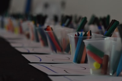 A row of plastic cups filled with colorful markers and crayons is placed on top of several sheets of paper. The scene appears to be part of an art or craft setup, possibly for a classroom or workshop.
