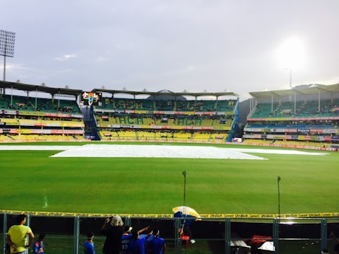 A cricket stadium with a covered pitch in the center, surrounded by vibrant green grass and multi-tiered seating areas. The stands are partially filled with spectators, and a group of people are visible in the foreground near a boundary fence. Floodlights illuminate the stadium as the sky appears overcast.