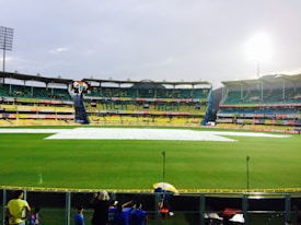 A cricket stadium with a covered pitch in the center, surrounded by vibrant green grass and multi-tiered seating areas. The stands are partially filled with spectators, and a group of people are visible in the foreground near a boundary fence. Floodlights illuminate the stadium as the sky appears overcast.