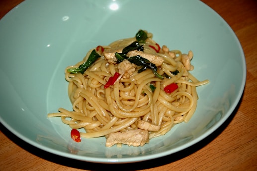 A bowl of noodles mixed with pieces of chicken, green leaves, and red chili peppers. The noodles are arranged in a spiral with visible seasoning on them, and the dish is presented in a light blue bowl on a wooden table.