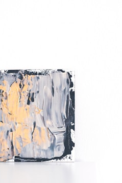 A piece of abstract art featuring thick brushstrokes in shades of black, gray, yellow, and white. The artwork occupies a corner of the frame against a white background, with a textured surface and dynamic patterns.