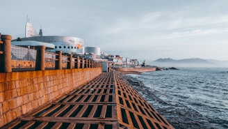 A coastal scene with a robust concrete and metal railing walkway overshadowing a rocky shoreline with gentle ocean waves. In the background, a modern, space-age building with circular and angular features stands alongside smaller structures. The distant mountains rise softly against a clear sky.