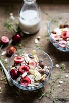 Two glass bowls containing a mixture of oats, nuts, and sliced red cherries are placed on a wooden table. A glass milk bottle partially filled with milk is in the background. Scattered around are loose oat flakes and greenery, creating a rustic breakfast setting.