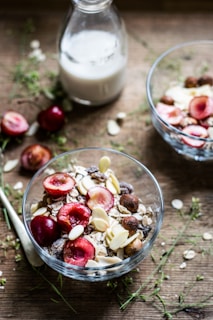 Two glass bowls containing a mixture of oats, nuts, and sliced red cherries are placed on a wooden table. A glass milk bottle partially filled with milk is in the background. Scattered around are loose oat flakes and greenery, creating a rustic breakfast setting.