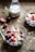 Two glass bowls containing a mixture of oats, nuts, and sliced red cherries are placed on a wooden table. A glass milk bottle partially filled with milk is in the background. Scattered around are loose oat flakes and greenery, creating a rustic breakfast setting.