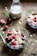 Two glass bowls containing a mixture of oats, nuts, and sliced red cherries are placed on a wooden table. A glass milk bottle partially filled with milk is in the background. Scattered around are loose oat flakes and greenery, creating a rustic breakfast setting.
