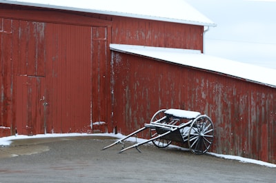 A rustic red wooden barn stands in the cold, with patches of snow clinging to its surface. An old wooden cart, also dusted with snow, is leaning against the barn. The ground is partially covered in snow, with a mix of dirt and gravel.