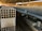 Rows of stacked concrete blocks are arranged neatly in an industrial factory setting with overhead metal beams and a concrete floor. The blocks have a hollow design and are organized along a long aisle. There are a few people visible in the distance, suggesting work activity.