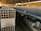 Rows of stacked concrete blocks are arranged neatly in an industrial factory setting with overhead metal beams and a concrete floor. The blocks have a hollow design and are organized along a long aisle. There are a few people visible in the distance, suggesting work activity.