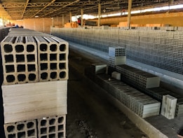 Rows of stacked concrete blocks are arranged neatly in an industrial factory setting with overhead metal beams and a concrete floor. The blocks have a hollow design and are organized along a long aisle. There are a few people visible in the distance, suggesting work activity.