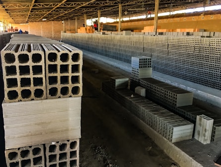 Rows of stacked concrete blocks are arranged neatly in an industrial factory setting with overhead metal beams and a concrete floor. The blocks have a hollow design and are organized along a long aisle. There are a few people visible in the distance, suggesting work activity.