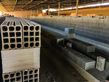 Rows of stacked concrete blocks are arranged neatly in an industrial factory setting with overhead metal beams and a concrete floor. The blocks have a hollow design and are organized along a long aisle. There are a few people visible in the distance, suggesting work activity.
