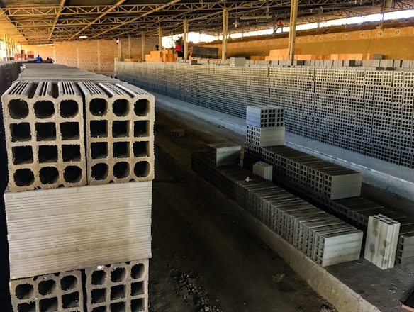 Rows of stacked concrete blocks are arranged neatly in an industrial factory setting with overhead metal beams and a concrete floor. The blocks have a hollow design and are organized along a long aisle. There are a few people visible in the distance, suggesting work activity.