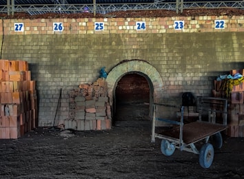 A brick kiln with numbered sections from 22 to 27 is visible. In the foreground, there are stacks of bricks on either side of a dark entrance. A cart with two wheels is positioned to the right, next to more bricks and some colorful fabric. The ground appears to be covered with a layer of dirt, and lighting is minimal, suggesting an industrial setting.