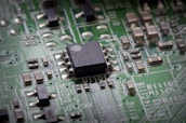 A close-up view of a green printed circuit board featuring a prominent black integrated circuit in the center. The board is populated with various small components such as resistors and capacitors, connected by silver traces. White alphanumeric markings are visible on the board's surface.