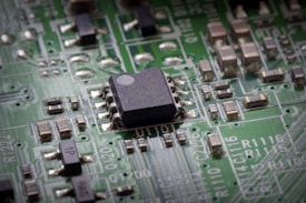 A close-up view of a green printed circuit board featuring a prominent black integrated circuit in the center. The board is populated with various small components such as resistors and capacitors, connected by silver traces. White alphanumeric markings are visible on the board's surface.