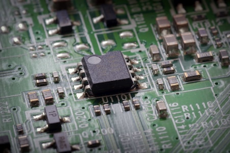 A close-up view of a green printed circuit board featuring a prominent black integrated circuit in the center. The board is populated with various small components such as resistors and capacitors, connected by silver traces. White alphanumeric markings are visible on the board's surface.
