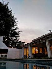 A tranquil outdoor scene featuring a house with glowing windows, indicating evening or early night. The house has a white exterior and a red-tiled roof, situated next to a pool reflecting the structure and the twilight sky. Tall, dark trees stand against the dimming light, providing a serene and quiet atmosphere.