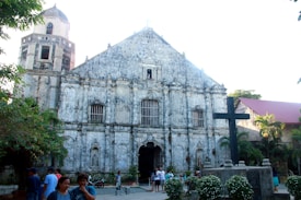 An old, rustic church with a stone facade featuring arched windows and a central entrance. A large black cross is prominently displayed in the foreground, surrounded by well-maintained foliage and a few flower bushes. Several people are gathered in the open courtyard area, engaging in casual conversation, with the atmosphere suggesting a community setting. The sky is clear, highlighting the architectural details of the building.