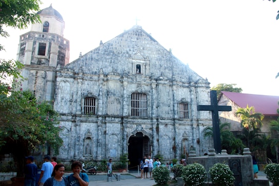 An old, rustic church with a stone facade featuring arched windows and a central entrance. A large black cross is prominently displayed in the foreground, surrounded by well-maintained foliage and a few flower bushes. Several people are gathered in the open courtyard area, engaging in casual conversation, with the atmosphere suggesting a community setting. The sky is clear, highlighting the architectural details of the building.