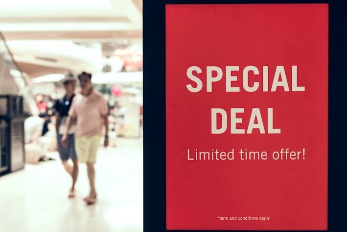A red sign displays the words 'SPECIAL DEAL' and 'Limited time offer!' in bold white letters, standing prominently in a shopping mall setting. In the background, two people walk past the sign, out of focus.