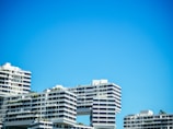 Several modern apartment buildings with a unique architectural design, featuring overhanging sections and geometric patterns, are set against a clear blue sky. The buildings have multiple balconies and large windows, with some greenery visible on the terraces.