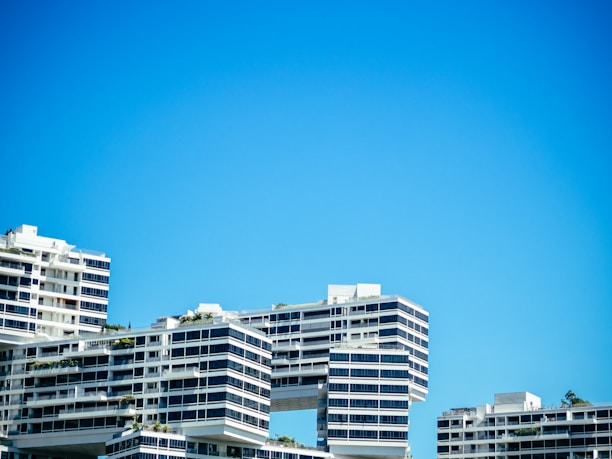 Several modern apartment buildings with a unique architectural design, featuring overhanging sections and geometric patterns, are set against a clear blue sky. The buildings have multiple balconies and large windows, with some greenery visible on the terraces.