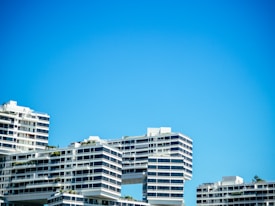 Several modern apartment buildings with a unique architectural design, featuring overhanging sections and geometric patterns, are set against a clear blue sky. The buildings have multiple balconies and large windows, with some greenery visible on the terraces.