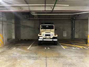 An old beige off-road vehicle is parked in an underground parking garage space 73. The garage has concrete walls and ceiling with visible pipes and electrical conduits. There are designated parking spaces marked with white and yellow lines on the ground, and a no parking warning sign is visible on the wall.