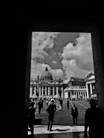 A black and white photograph capturing a view of a grand architectural structure with a prominent dome, likely St. Peter's Basilica, set against a sky filled with scattered clouds. Numerous people are walking towards the basilica, with the scene framed by the dark silhouette of an archway.