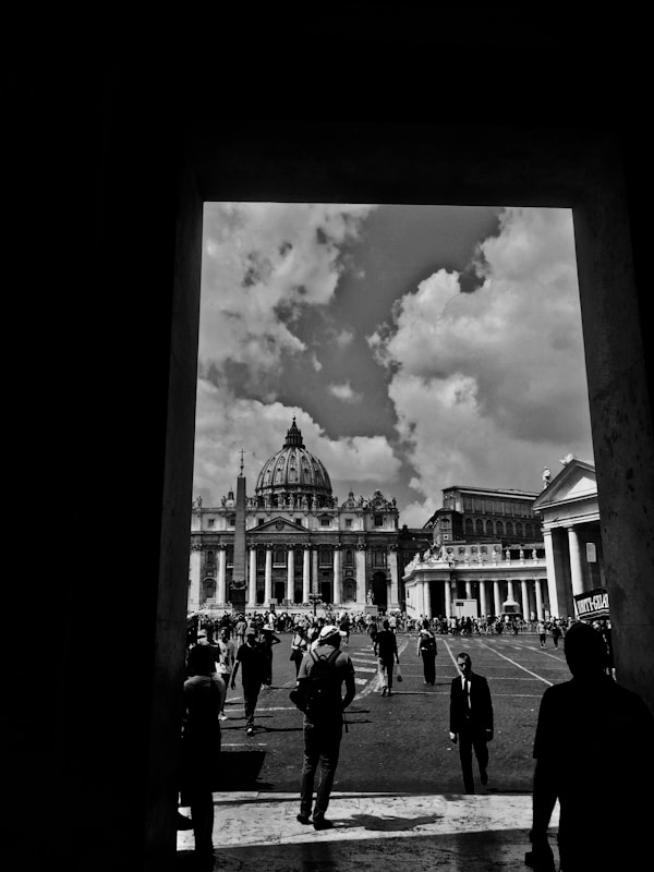A black and white photograph capturing a view of a grand architectural structure with a prominent dome, likely St. Peter's Basilica, set against a sky filled with scattered clouds. Numerous people are walking towards the basilica, with the scene framed by the dark silhouette of an archway.