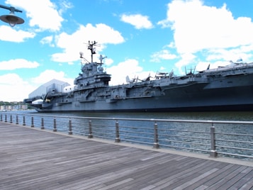 A large aircraft carrier is docked near a wooden boardwalk, with a partly cloudy sky above and city buildings visible in the distance. The ship is massive and detailed, with various structures and equipment on deck.