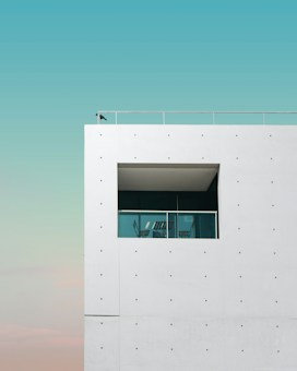 A modern minimalist building with a white facade featuring a rectangular window. The window frames a glass balcony with metal railings and a couple of chairs. A single bird is perched on the corner of the building. The sky is a clear blue with a gradient towards the horizon.
