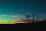 A wire fence is silhouetted against a colorful sunset sky, featuring shades of teal, pink, and orange. The fence leans slightly at an angle and is set against a dark, flat landscape.
