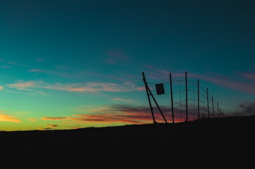 A wire fence is silhouetted against a colorful sunset sky, featuring shades of teal, pink, and orange. The fence leans slightly at an angle and is set against a dark, flat landscape.