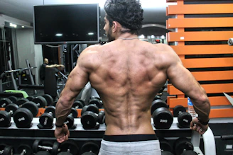 A muscular person with a well-defined back stands in front of a rack of dumbbells in a gym. The individual’s back muscles are prominently displayed, indicating strength and fitness. In the background, gym equipment is visible along with another person seated.