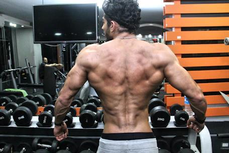A muscular person with a well-defined back stands in front of a rack of dumbbells in a gym. The individual’s back muscles are prominently displayed, indicating strength and fitness. In the background, gym equipment is visible along with another person seated.