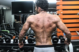 A muscular person with a well-defined back stands in front of a rack of dumbbells in a gym. The individual&rsquo;s back muscles are prominently displayed, indicating strength and fitness. In the background, gym equipment is visible along with another person seated.