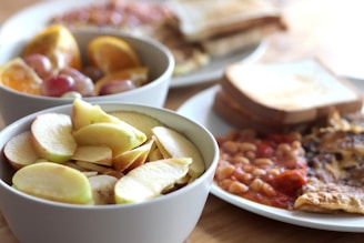 A table is set with bowls and plates of various breakfast foods, including sliced apples, oranges, grapes, toast, and a dish with baked beans and an omelette.